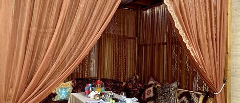 A traditional outdoor seating area with a table set for dining, surrounded by patterned cushions and a decorative canopy.
