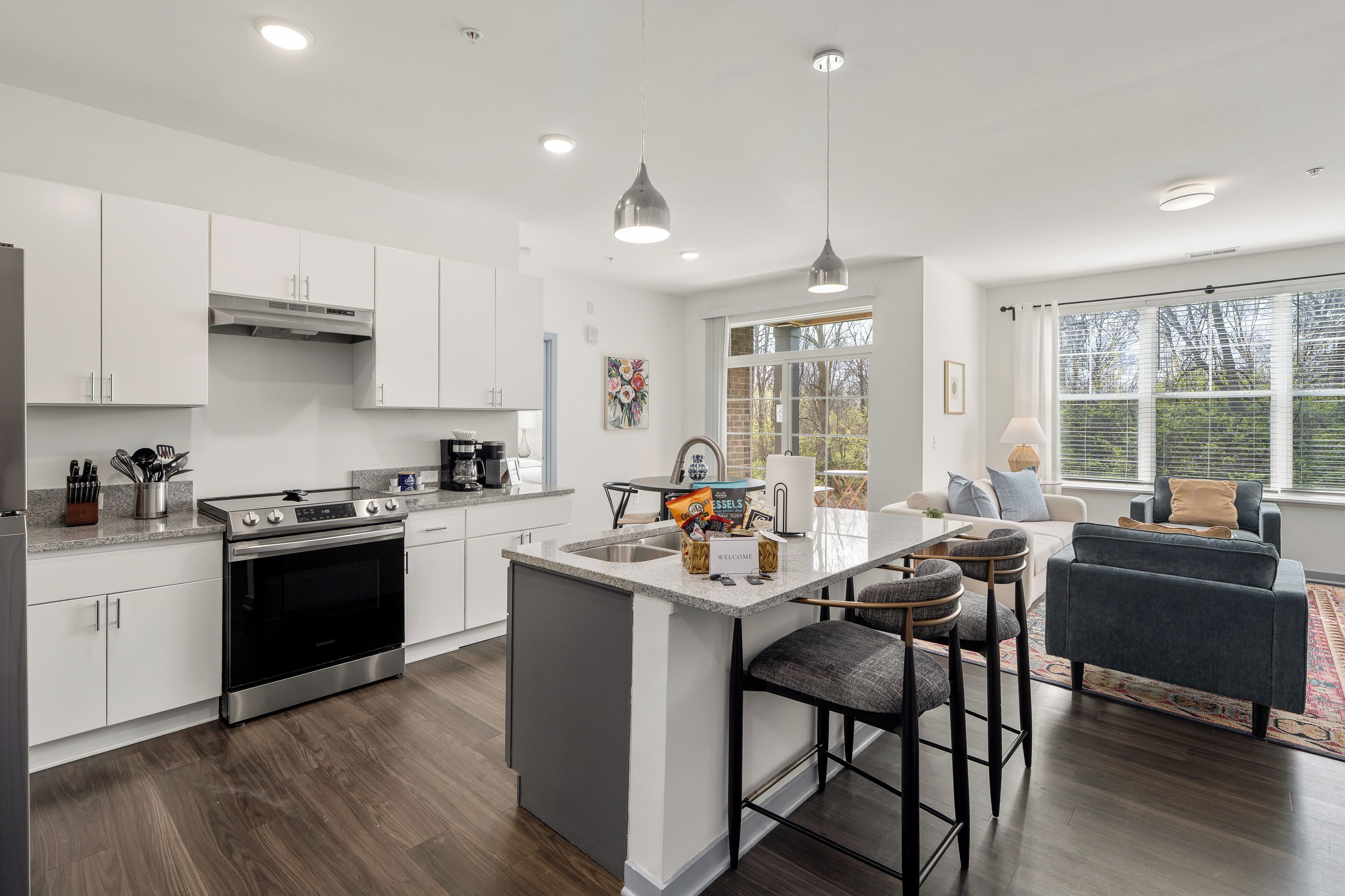 A modern kitchen with white cabinets, a central island, bar stools, and a dining area with a sofa and table.