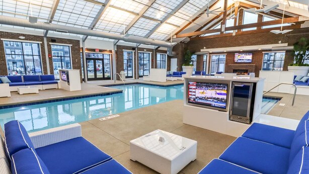 An indoor swimming pool area with blue lounge chairs, a white coffee table, and a TV screen displaying a sports event. - Dublin 1BR | Pets OK | King bed | 0 stairs (Dublin)