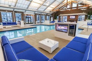Et indendørs swimmingpoolområde med blå liggestole, et hvidt sofabord og et tv, der viser et sportsarrangement.