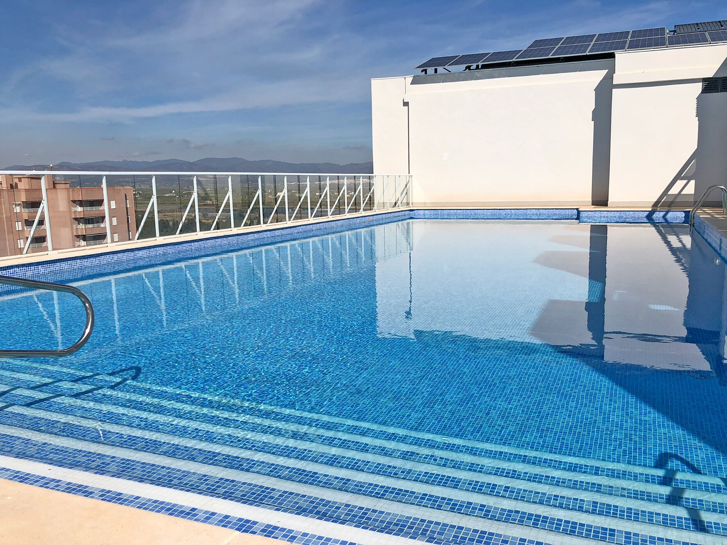 A rooftop swimming pool with clear blue water, a metal ladder, and solar panels on the roof.