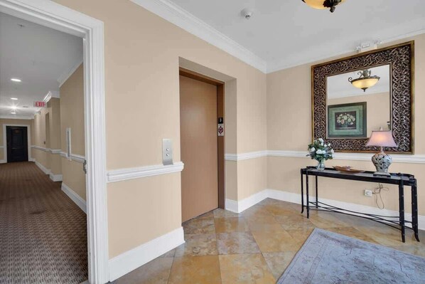 Eine Hotellobby mit einer beige gestrichenen Wand, einer Holztür, einem schwarzen Konsolentisch mit einer Lampe und einem gemusterten Teppich.
