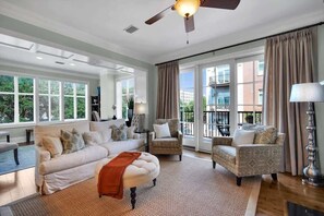Ein geräumiges Wohnzimmer mit einem weißen Sofa, zwei gemusterten Sesseln, einem runden Hocker und einem Deckenventilator. Große Fenster bieten einen Blick ins Freie.