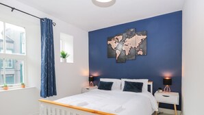 Un dormitorio con cama blanca, pared azul como detalle, un cuadro de mapa mundial, dos mesitas de noche con lámparas y una ventana con cortina.