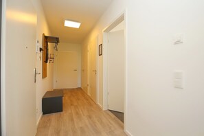A narrow hallway with wooden flooring, white walls, and a ceiling light. There are doors on both sides, one of which has a small black object on the floor.