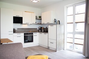 A modern kitchen with white cabinets, a built-in oven, and a microwave. There is a dining area with a table and chairs, and a large window providing natural light.