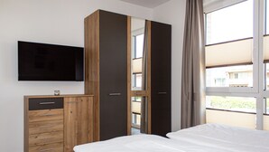 A bedroom with a bed, a TV mounted on the wall, a wardrobe, and a window with a view of buildings outside.