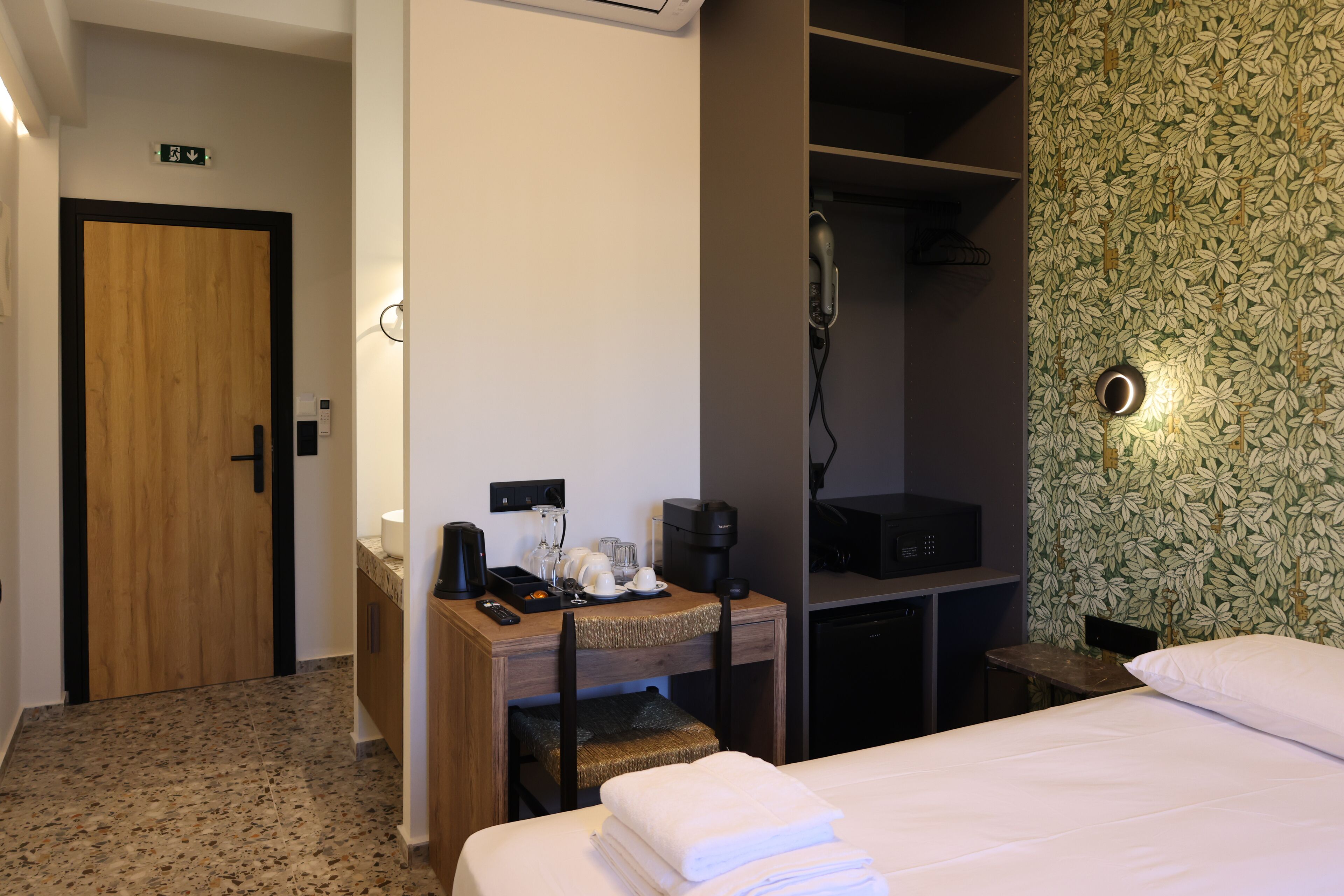 Habitación de hotel con puerta de madera, un escritorio con cafetera, teléfono y cama con sábanas blancas.