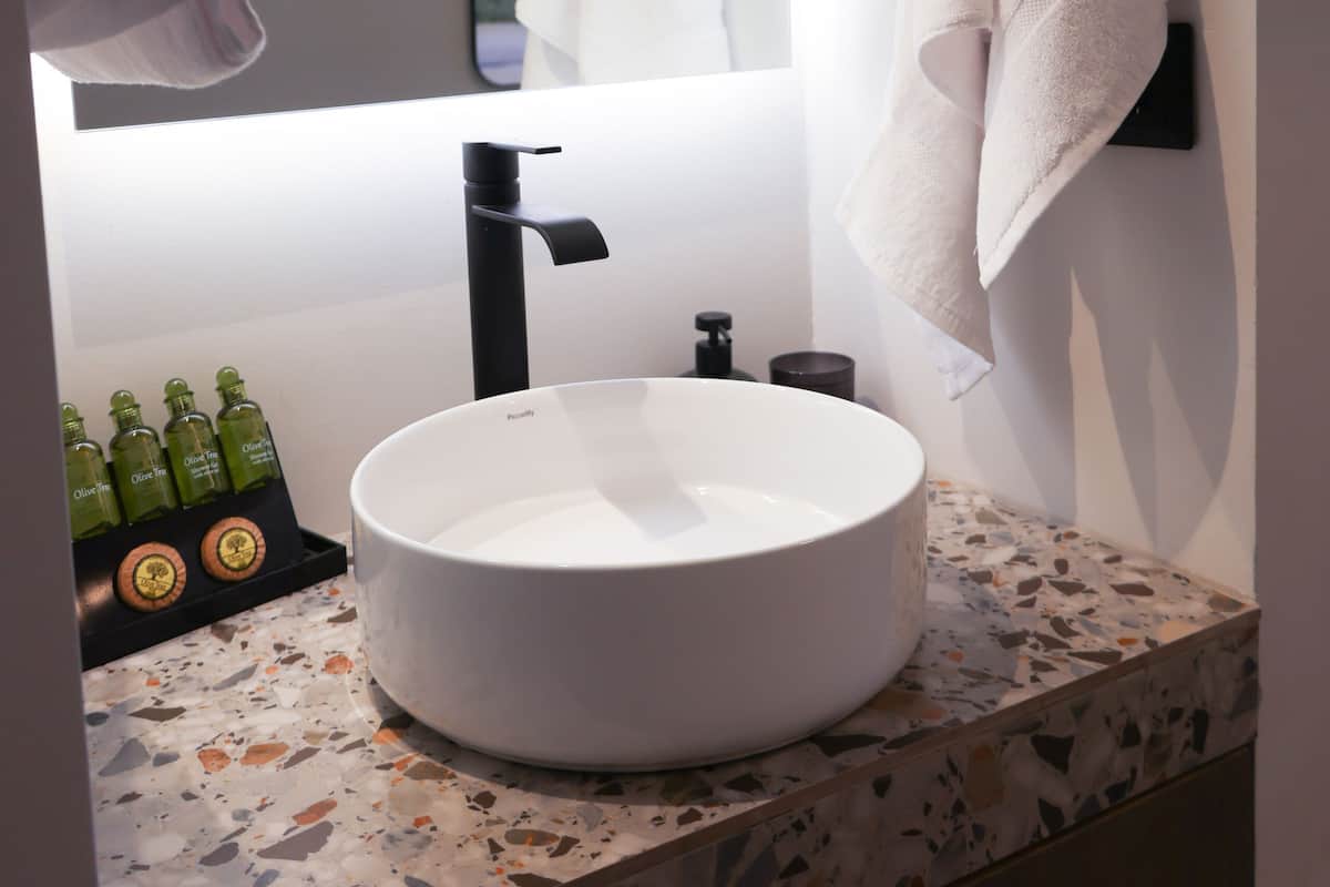 Een badkamer met een rond wit wastafeltje, een zwarte kraan en een bezaaid aanrecht. Links staan groene flesjes en rechts een handdoekhouder.