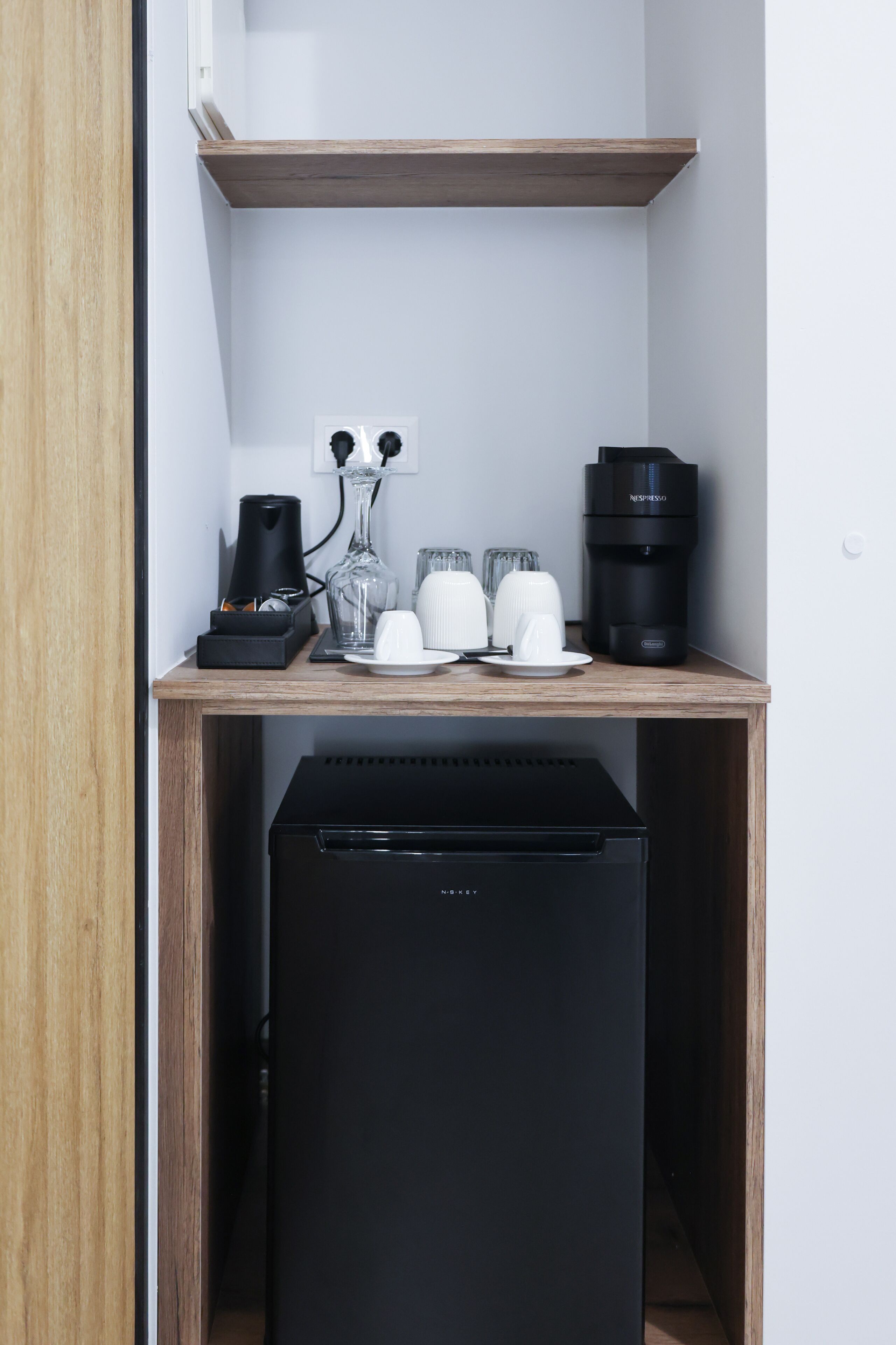 Een kleine keuken met een koffiezetapparaat, een Nespresso machine en een zwarte koelkast.