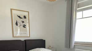 A double bed with white linens, a framed floral artwork on the wall, a bedside table with a lamp, and a window with curtains. - Strandnahe 2-zimmer-fewo für 3 Personen mit Wlan (Graal-Müritz)
