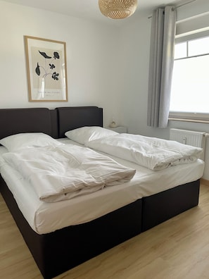 A double bed with white linens, a framed floral artwork on the wall, a bedside table with a lamp, and a window with curtains. - Strandnahe 2-zimmer-fewo für 3 Personen mit Wlan (Graal-Müritz)
