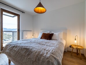 A bedroom with a large bed, a wooden floor, a window with a view of mountains, and a hanging light fixture.
