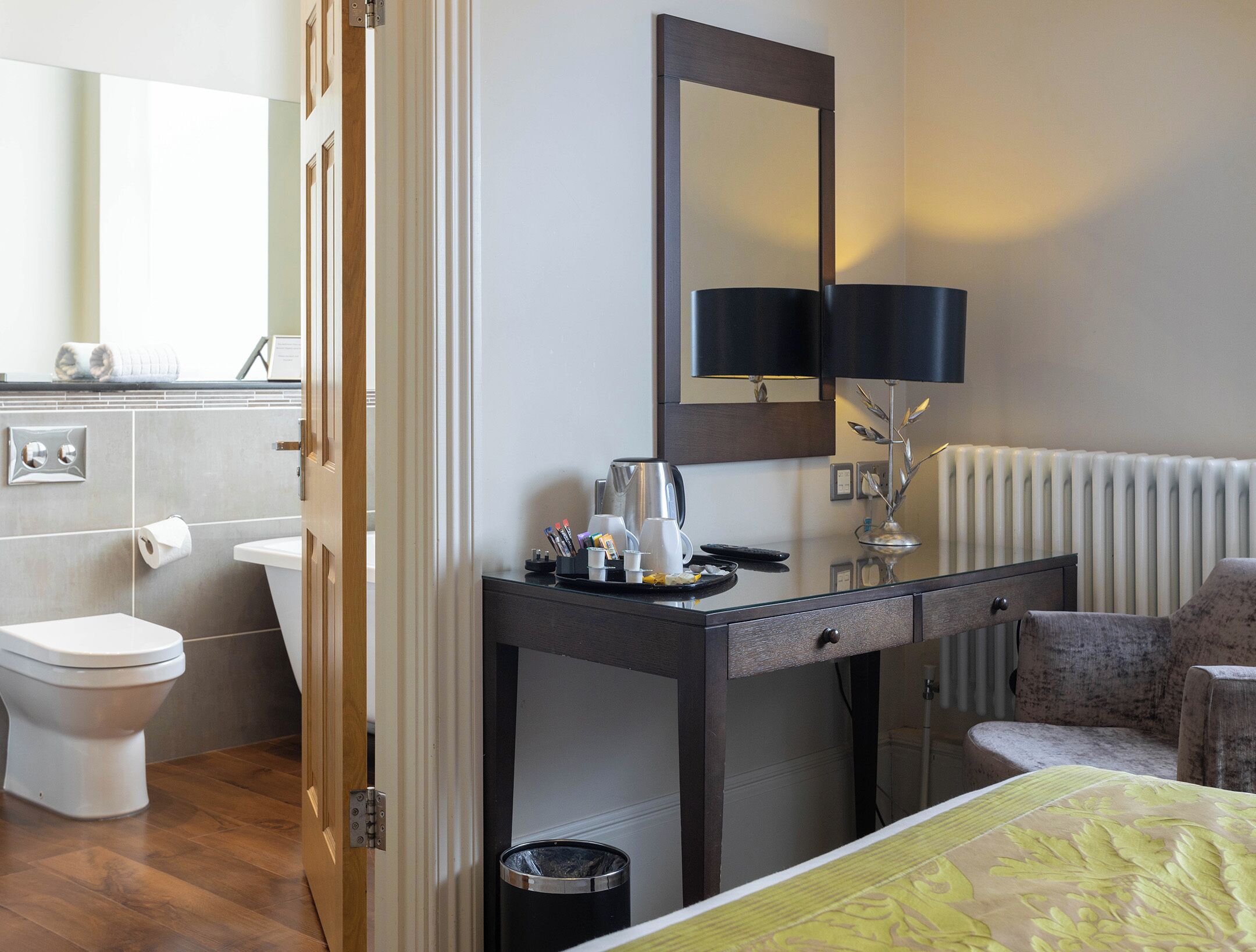 A hotel room with a wooden floor, a desk with a lamp, a chair, a bed with a yellow bedspread, a toilet, and a mirror on the wall.