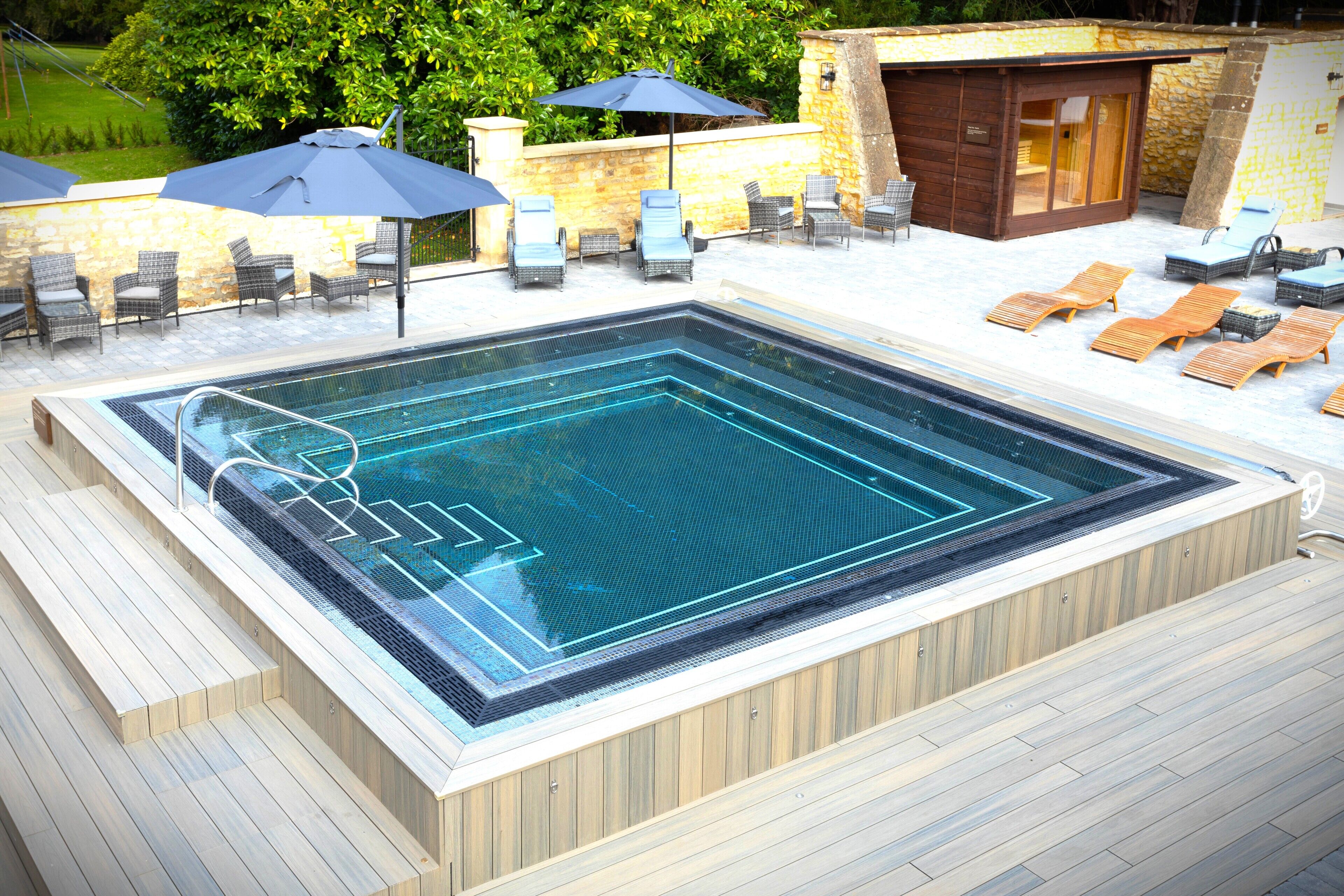 Een rechthoekig jacuzzi met een houten terras eromheen, voorzien van lounge fauteuils en parasols.