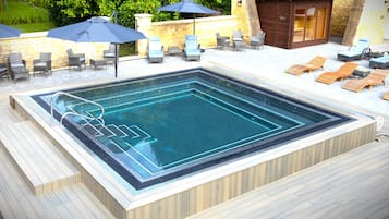 A rectangular hot tub with a wooden deck surrounding it, equipped with lounge chairs and umbrellas.