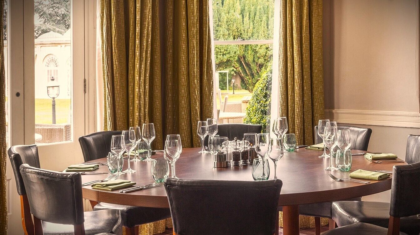 A round dining table set for six with chairs, a window with curtains, and a view of greenery outside.