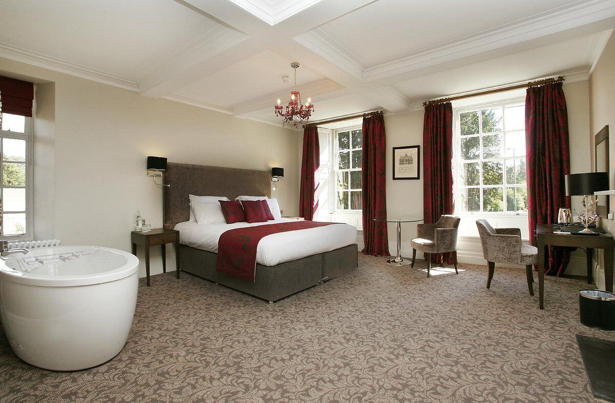A bedroom with a large bed, a sitting area with two chairs, a side table, a large white bathtub, and a window with red curtains.