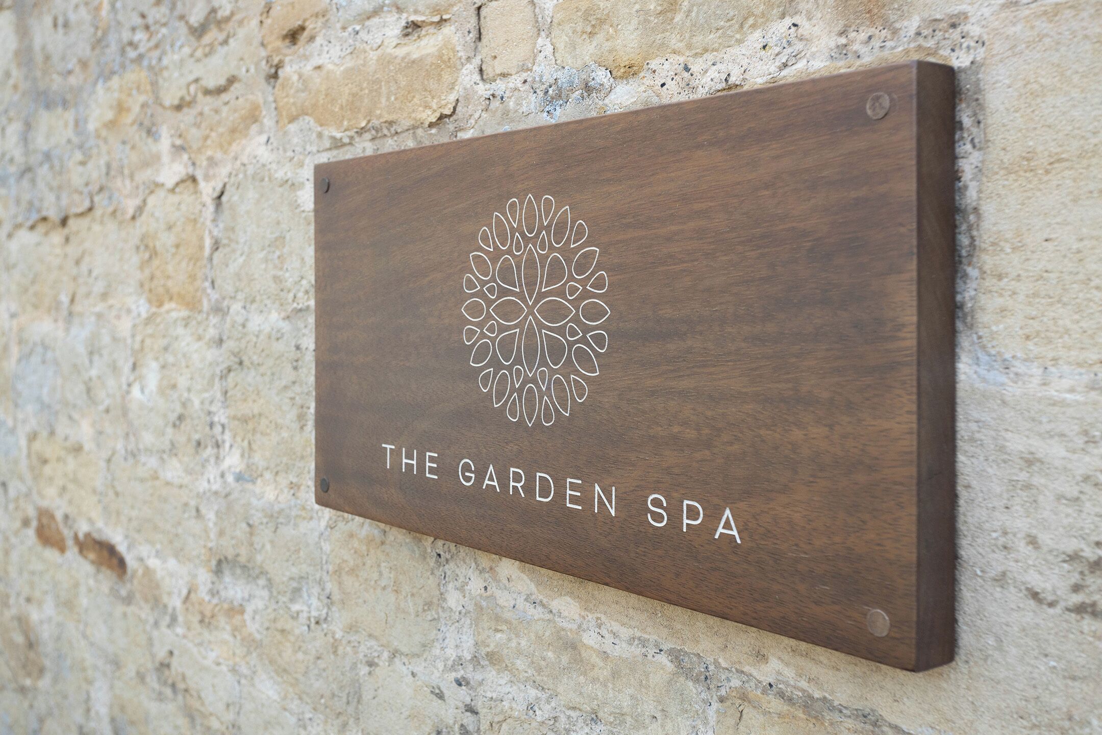 A wooden sign with a floral design and the text "The Garden Spa" mounted on a stone wall.
