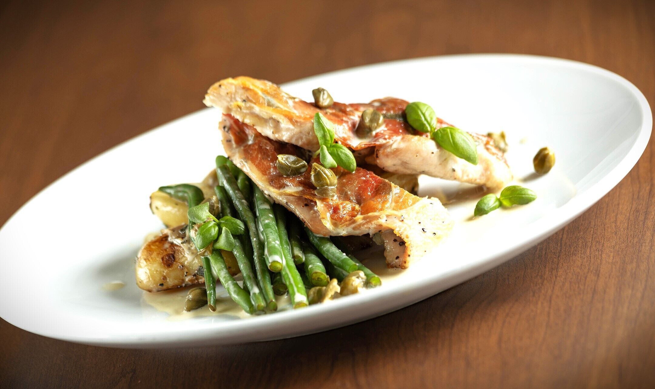 Une assiette blanche contenant du poulet rôti, des haricots verts et des câpres, posée sur une table en bois.