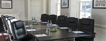 Un salón de conferencias con una larga mesa, sillas de cuero negro, botellas de agua, vasos y un plato de manzanas.