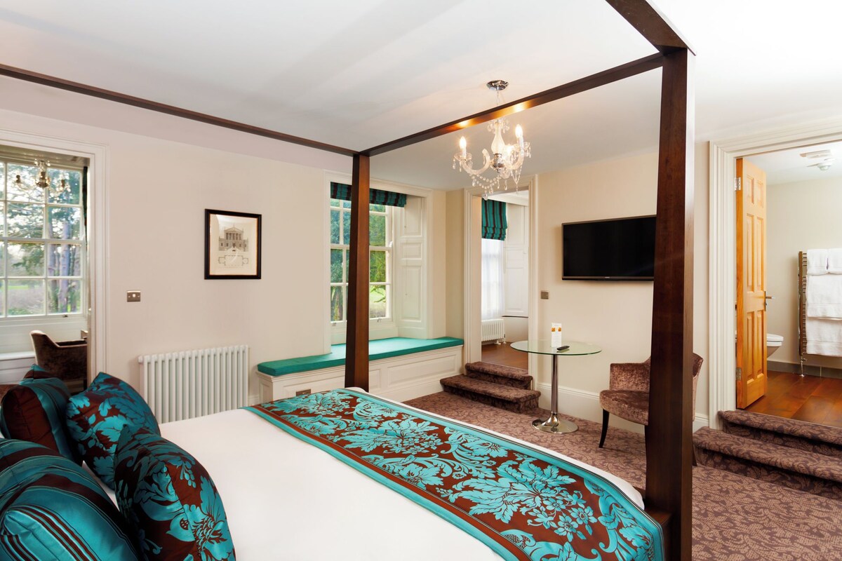 Una cama con dosel con colcha estampada, un candelabro, vista al exterior a través de una ventana, un televisor y un baño con toallero.