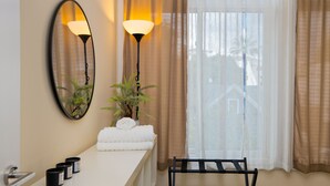 Ein Badezimmer mit einem weißen Waschbecken, zwei Wandleuchten, einem Spiegel, einer Topfpflanze, einem Handtuchhalter und einem Fenster mit durchsichtigen Vorhängen.