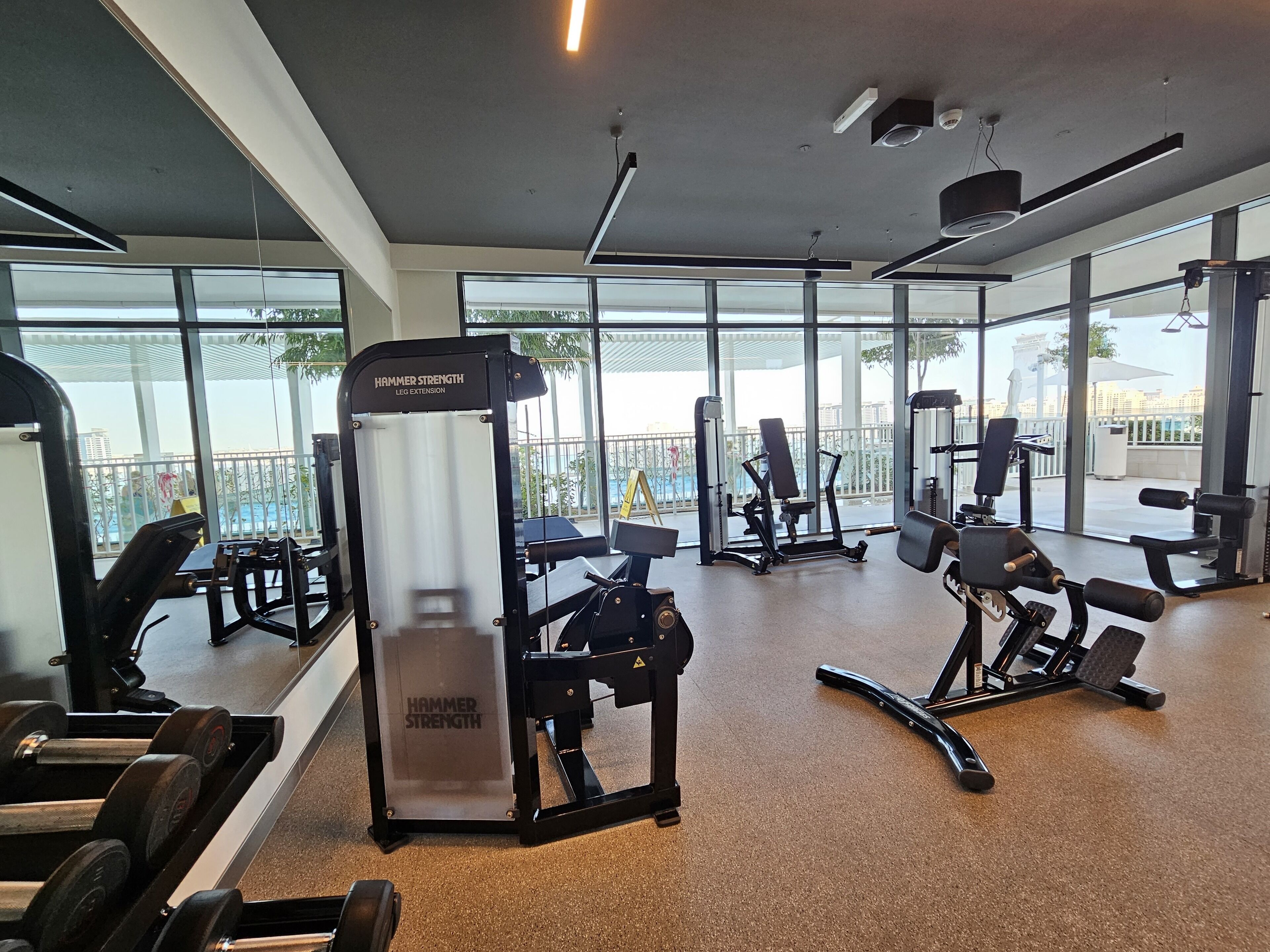 Ein Fitnessstudio mit unterschiedlichen Trainingsgeräten, großen Fenstern und Blick auf die Stadtlandschaft.