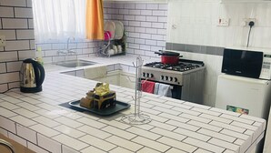 Une cuisine avec un mur recouvert de carreaux blancs, une cuisinière, un évier et un réfrigérateur. On y trouve deux chaises bleues et un petit îlot central avec une bouilloire et une théière.