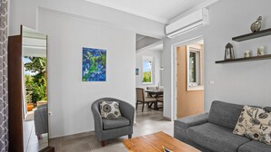 Una sala de estar moderna con un sofá gris, una mesa de centro de madera y un sillón gris con un cojín estampado. Un cuadro abstracto azul cuelga en la pared, y se aprecia un comedor a través de una puerta abierta.