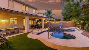 Patio trasero residencial con piscina, mobiliario exterior e iluminación con luces colgantes.