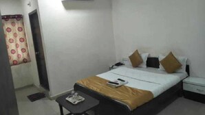 A hotel room with a bed, bedside table, nightstand, and an air conditioning unit. - Hotel Silver Inn - Naroda (Ahmedabad)