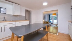 Eine moderne Küche mit weißen Schränken, einer grauen Arbeitsplatte und einer hölzernen Bank. Ein Essbereich mit Tisch und Stühlen bietet einen Blick ins Freie durch eine Glastür.