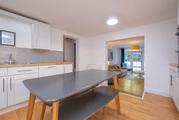 A modern kitchen with white cabinets, a grey countertop, and a wooden bench. There is a dining area with a table and chairs, and a view of the outdoors through a glass door.