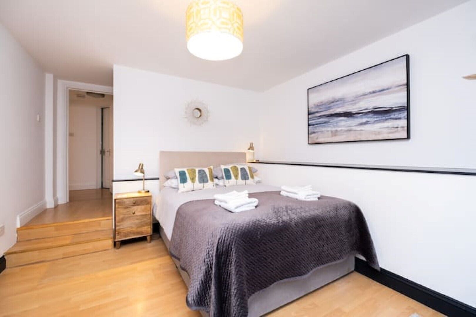 A modern hotel room with a wooden floor, a bed with a grey blanket, a nightstand with a lamp, and a painting on the wall.