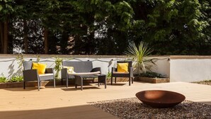 Eine Terrasse im Freien mit Korbmöbeln, einer Feuerstelle in der Mitte und einer weißen Wand im Hintergrund.