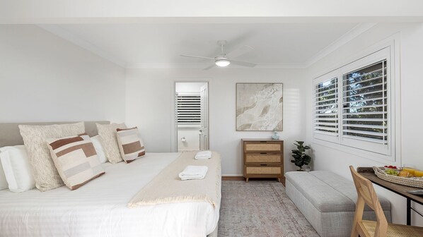Quarto com cama, mesas de cabeceira, ventoinha no teto, janela com persianas, obra de arte na parede e um pequeno espaço de estar.