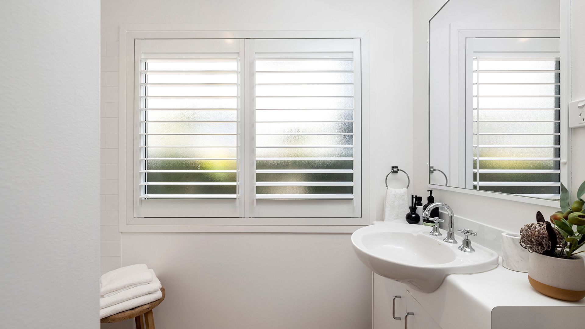 Une salle de bain avec un lavabo blanc, un miroir et une fenêtre avec des stores.