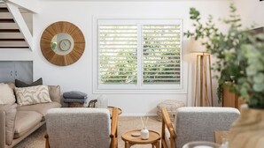 A living room with a sofa, two armchairs, a coffee table, a side table, a floor lamp, a mirror, and a window with blinds. - Cabane Sur Mer – views of Shoal Bay (Shoal Bay)