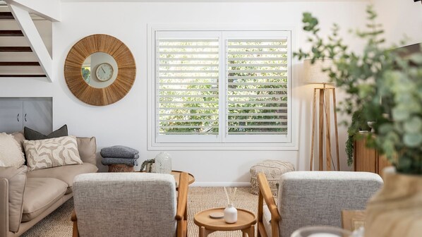 Una sala de estar con un sofá, dos sillones, una mesa de centro, una mesita auxiliar, una lámpara de pie, un espejo y una ventana con persianas.