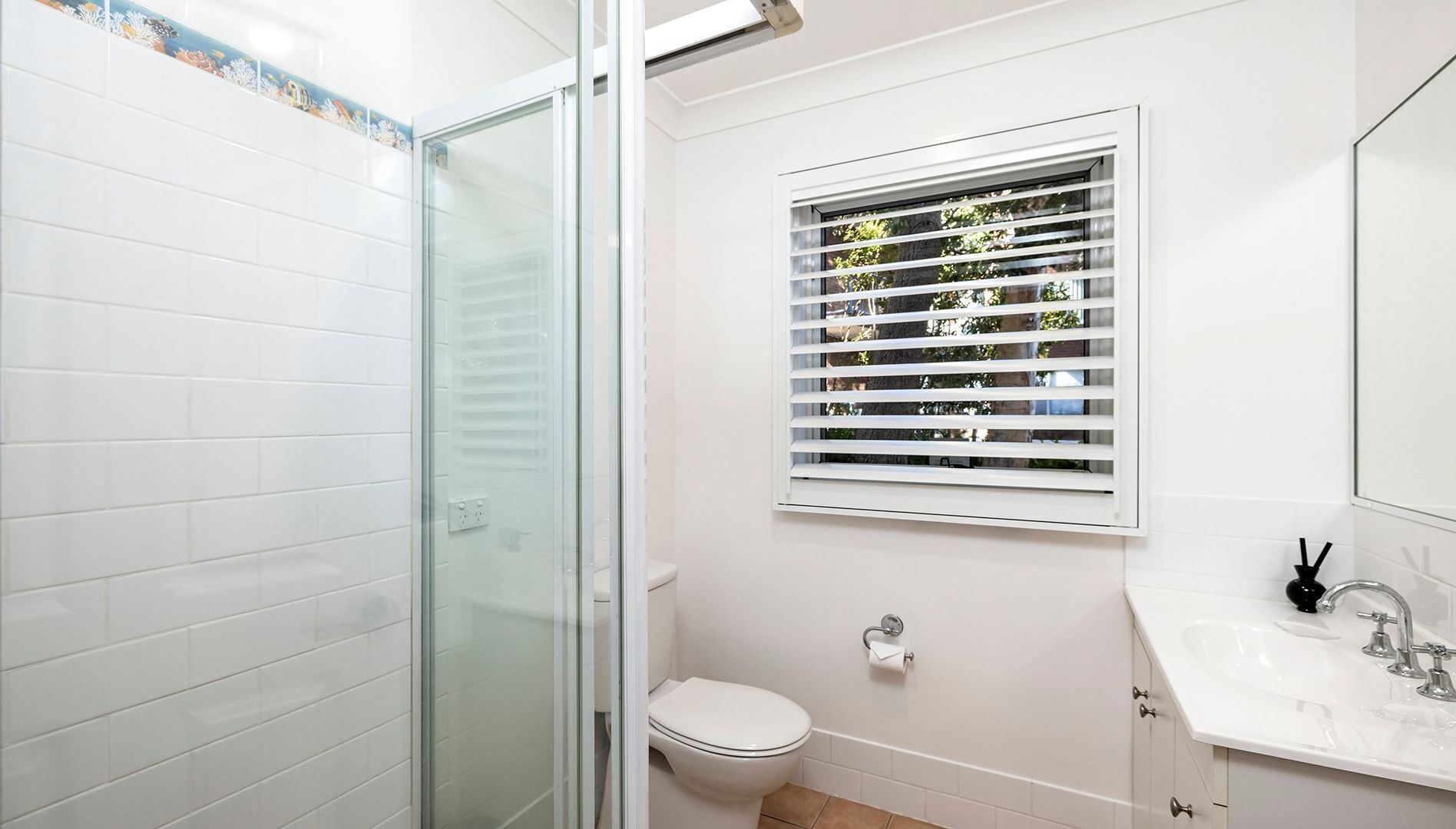 Une salle de bain avec une douche à parois de verre, des toilettes, une fenêtre avec des stores et un lavabo avec un miroir.