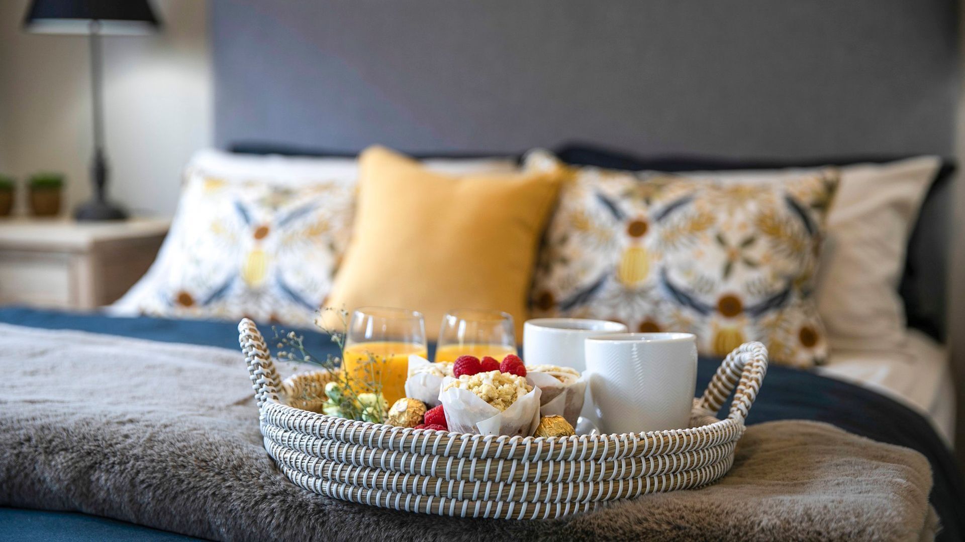 A wicker tray with breakfast items on a bed, including a cup, a glass of orange juice, and some pastries.