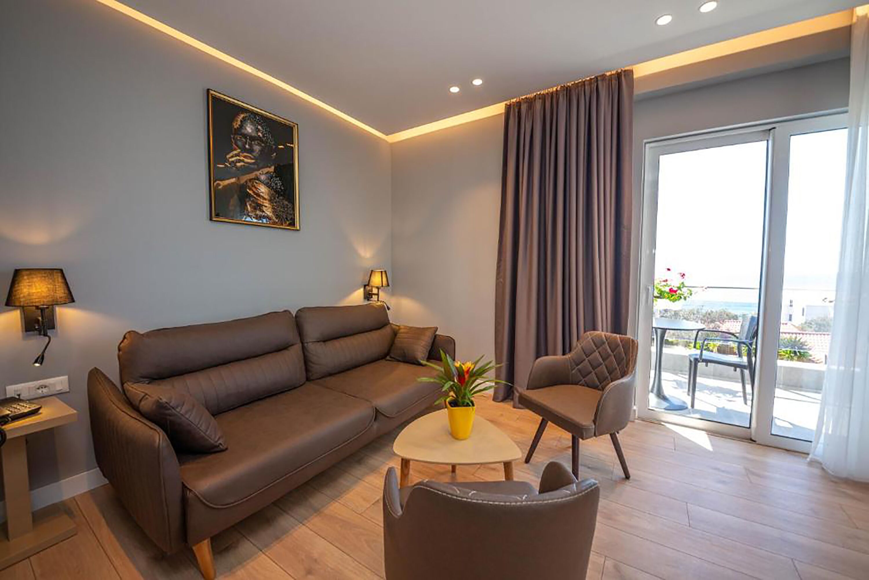 En moderne stue med en brun lædersofa, et rundt sofabord med en gul blomstervase, en polstret lænestol og et stort vindue med havudsigt.