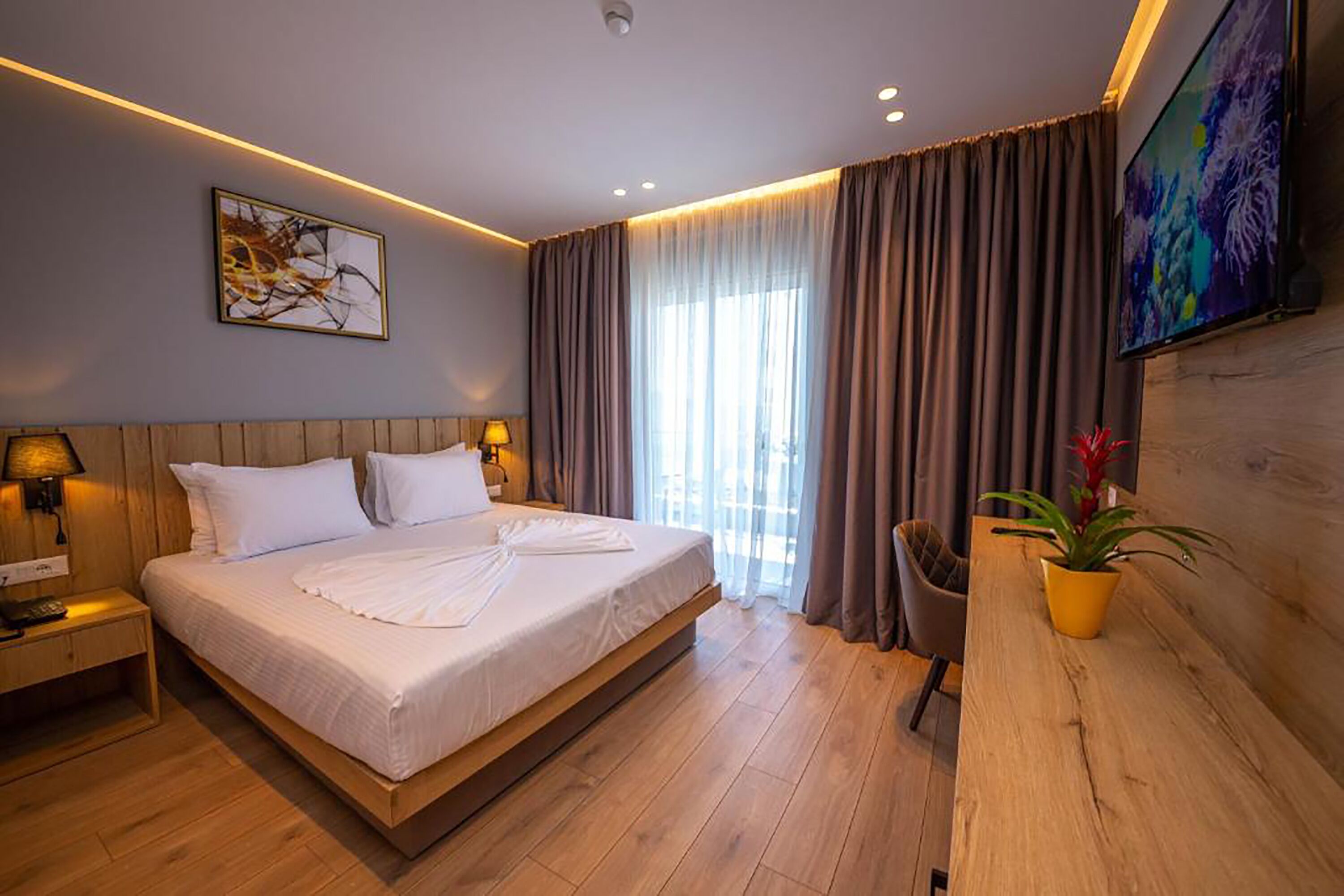 A modern hotel room with a large bed, wooden headboard, bedside lamps, a flat-screen TV, a wooden desk, and a potted plant.
