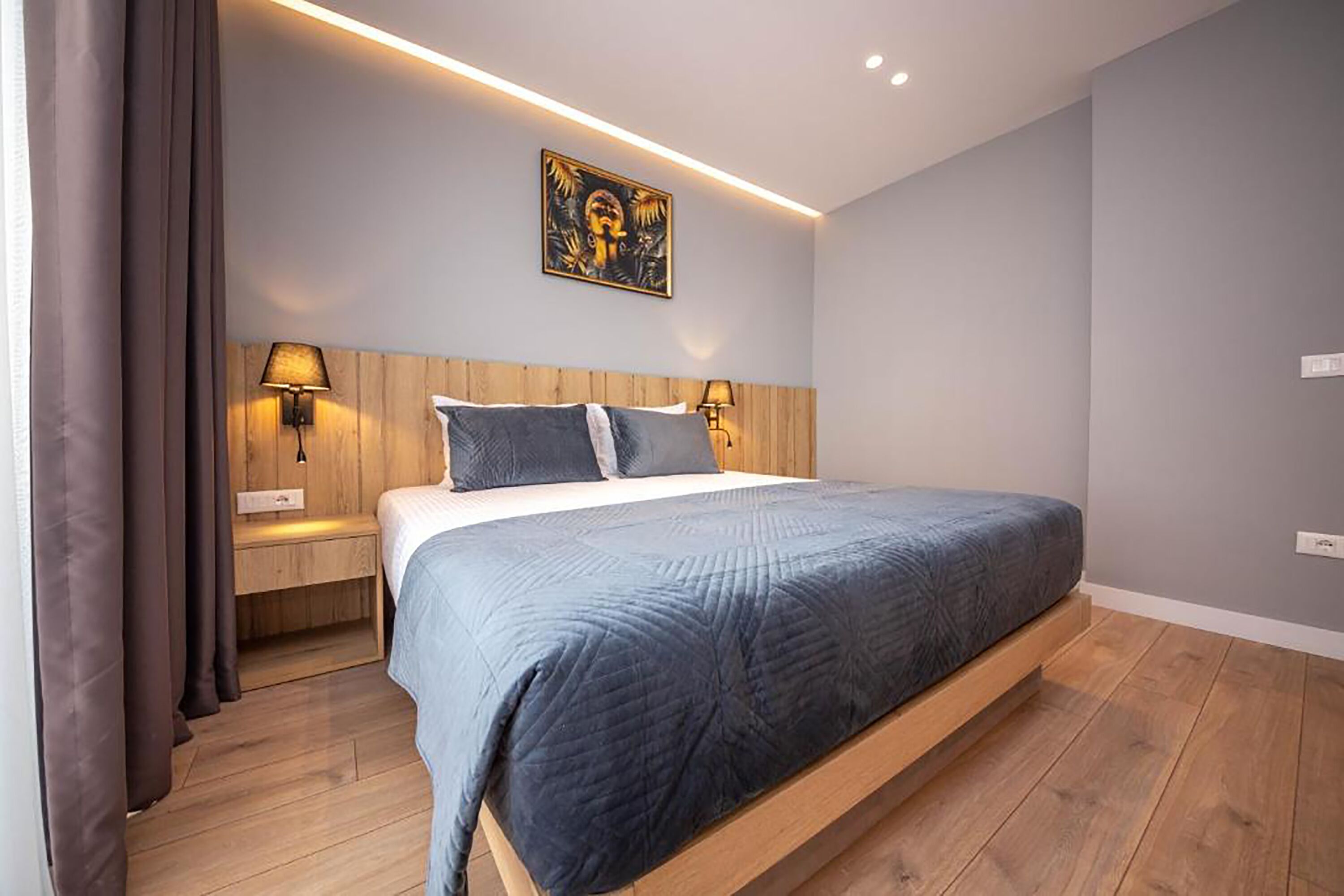 Un dormitorio moderno con cama, cabecero de madera, dos lámparas de noche, una mesita de noche y una obra de arte en la pared.