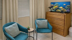 A room with two teal armchairs, a small round table with a plant, a wooden cabinet, and a television displaying an underwater scene. - 3-Suite Group Stay | Steps to Beach | Sleeps 13 | Save 15% if Book Non-Refundable (West Bay)