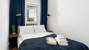 Un lit bien fait, avec du linge de lit blanc, une couverture bleu foncé et un bouquet de fleurs blanches. Il y a deux tables de chevet avec des lampes, et une fenêtre avec des rideaux bleus.