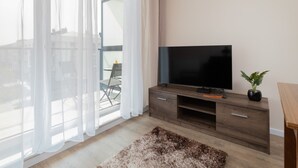 A modern living room with a flat-screen TV on a wooden TV stand, a large window with sheer curtains, a fluffy rug, and a potted plant. - Elegant Apartment in Gdynia | Balcony and Private Parking (Gdynia)