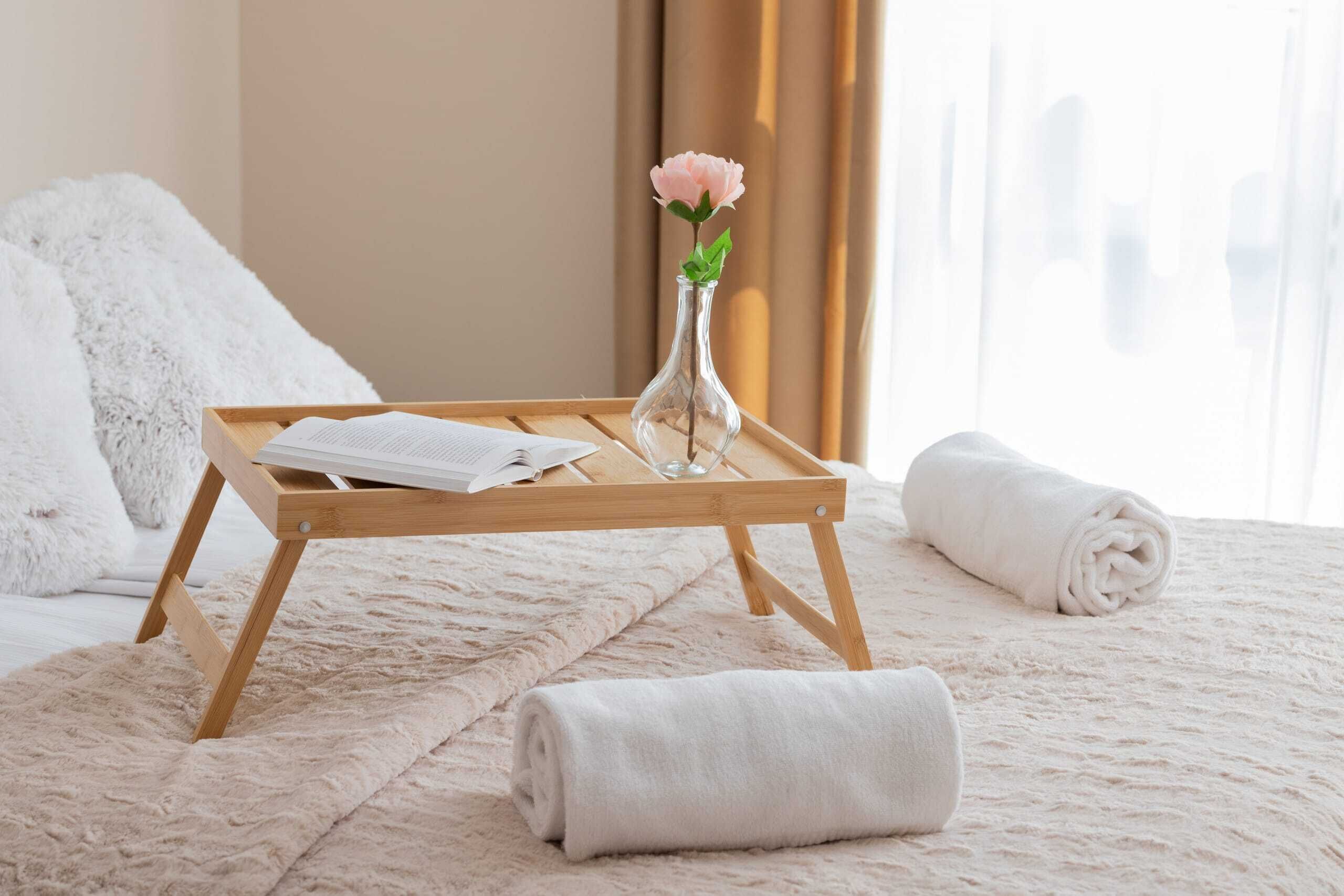 Un letto rifatto con cura, con una soffice coperta bianca, un vassoio di legno con un asciugamano arrotolato e un vaso con un fiore, e una finestra con tende.