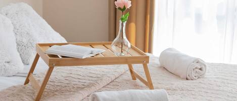 A neatly made bed with a fluffy white blanket, a wooden tray with a rolled towel and a vase with a flower, and a window with curtains.